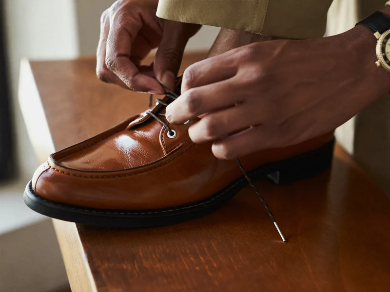 Mains ajustant les lacets d'une chaussure en cuir sur un banc d'essayage en bois.